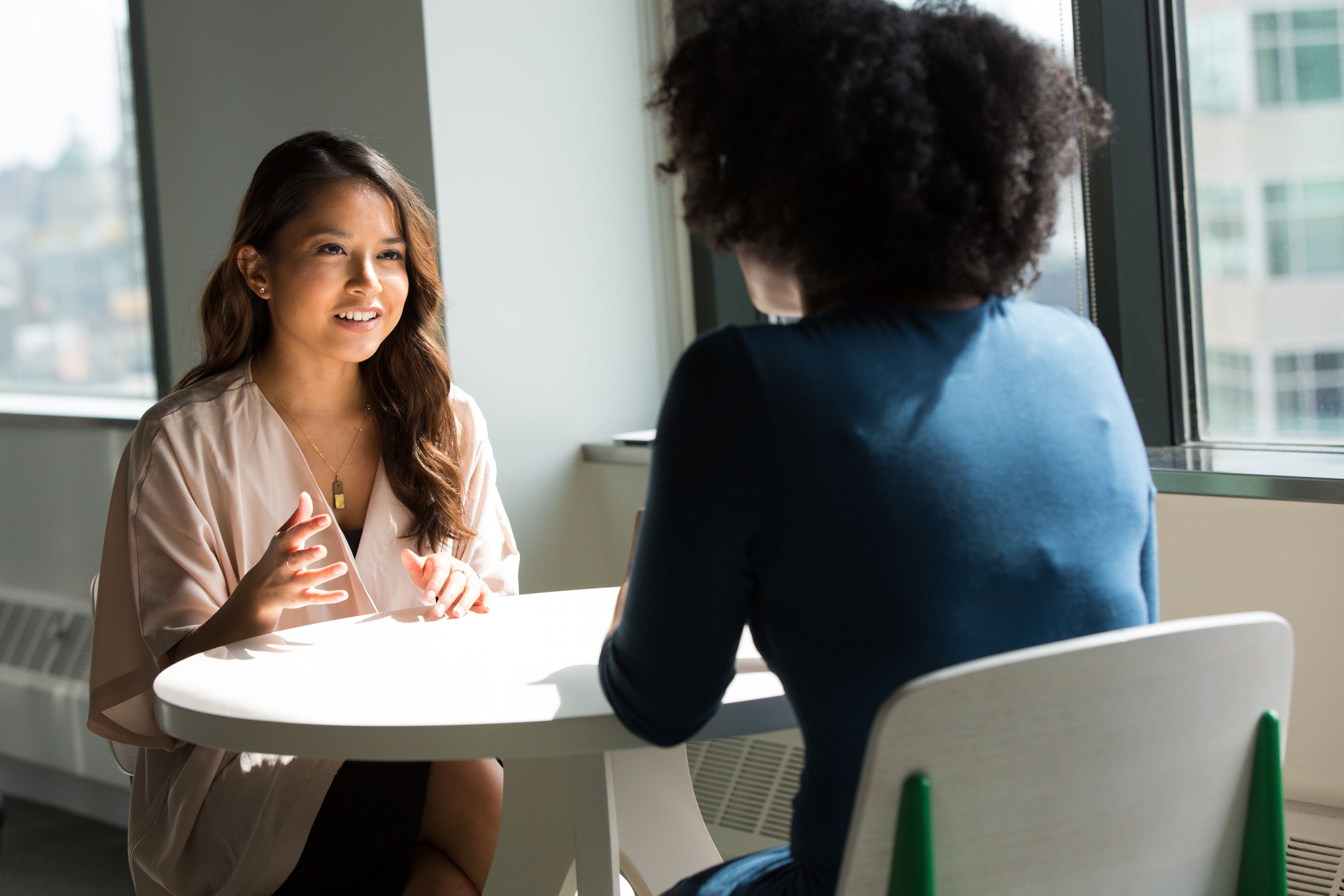 Two people talking across a round table by tall windows